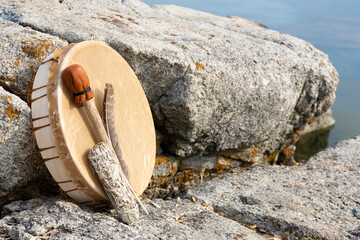 An image of a hand made leather meditation drum leaning on a rock face with sacred feather and sage smudge stick.