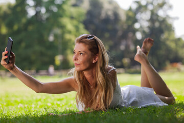 A cheerful young woman lies on the grass in a sunny park, capturing a selfie with her smartphone, embodying a moment of joy and relaxation.