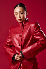 Wall Mural - Fashion asian brunette woman in red leather jacket posing with hands on hips and smiling against red background