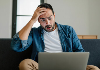 a man in casual attire reacts with shock while using a laptop in a modern living room