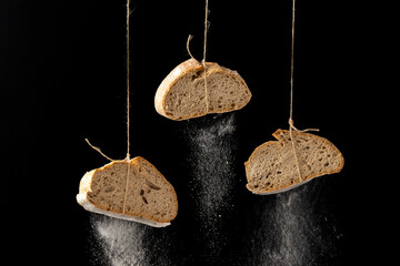 Three slices of rustic bread are suspended on strings with flour artfully falling against a dark background, creating a striking contrast and an abstract visual effect.