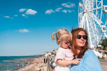 A mother holds her baby daughter next to a Ferris wheel against a blue sky background. Zelenogradsk, Russia - 20 June 2024