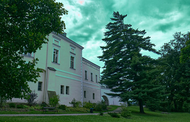 Photo of the castle park. Castle building with a light facade. A park with benches, trees and a castle will be under an overcast sky with clouds. Rainy day, cloudy.