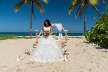 Beautiful wedding couple bride in lace wedding dress with bouquet of roses and groom reach their hands to sun near white arch on the beach of caribbean sea	