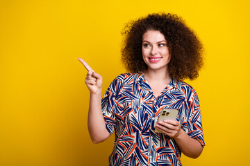 Poster - Photo portrait of pretty young girl hold device point empty space wear trendy colorful outfit isolated on yellow color background