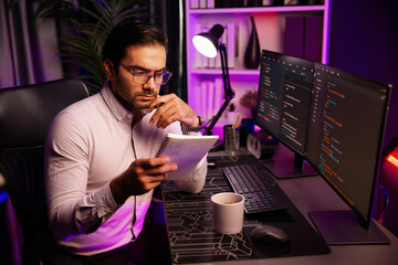 Smart IT developer working on software development coding on pc screen and reading note of newest application project, thinking program update online website data at neon light cyber office. Surmise.