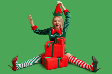 Wall Mural - Emotional young woman in elf costume with Christmas gift boxes on green background