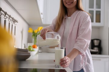 Poster - Young woman pouring milk from gallon bottle into glass at light countertop in kitchen, closeup