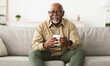© Prostock-studio - Mature Black Man Drinking Coffee Watching TV Sitting On Sofa And Smiling To Camera Holding Cup At Home, Wearing Eyeglasses. Retirement Lifestyle And Leisure Concept