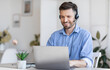 © Prostock-studio - Call center operator at work. Positive male manager wearing headset and typing on laptop at workplace in modern office, consulting clients