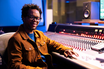 Fototapete - Portrait of african american sound engineer producing records in studio, working with digital audio software and professional mixing console in control room. Expert adjusting volume levels on songs.