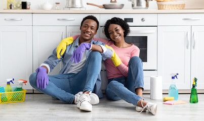 Tired black couple in rubber gloves sitting on floor next to stove among house-keeping supplies in cozy kitchen, hugging, resting after cleaning apartment, smiling at camera, copy space