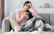 © Prostock-studio - Despaired sad young hindu lady with plaid, suffer from flu, sits on sofa and looks at thermometer in living room interior. Cold, fever, disease treatment at home and covid-19 lockdown, free space