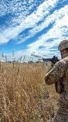 A soldier in camouflage gear holds a rifle and looks out over a field with tall dry grass.