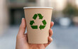 © Pongsatorn - Hand holding a recyclable paper cup with a green recycling symbol, promoting eco-friendly practices.