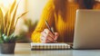 © Intelligent Horizons - Focused woman sitting at a sunlit desk writing in a notebook while referencing her laptop computer surrounded by a tidy and organized workspace with plants and other office essentials