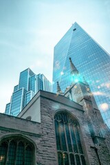 Poster - An old stone church building with a large arched window stands against a backdrop of modern glass skyscrapers in a large city.