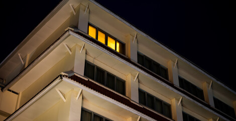 A tall building with a window that is lit up. The building is white and has a red trim