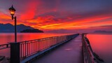 Fototapeta Pomosty - A long pier extends out into a calm lake with a dramatic red sunset. The pier is illuminated by a street lamp.