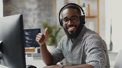 A young African American man wearing glasses and headphones, sitting at a computer, smiles and gives a thumbs up.