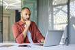 © Liubomir - Thoughtful man with laptop in modern office. Professional analyzing work strategy. Wearing casual attire, promoting focused approach to tasks. Phone and notebook nearby for efficiency.