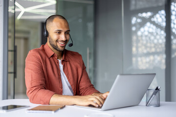 Wall Mural - Smiling professional wearing headset works on laptop in modern office setting. Engaged in online meeting using phone technology. Notebook and pen on table indicate readiness and focus.