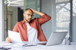 © Liubomir - Worried man in office holding document while looking at laptop with stressed expression. Professional setting suggests business concern, stress, and decision-making scenario.