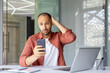 © Liubomir - Concerned man holds smartphone and looks worried while working in office. Laptop and notebook on desk suggest business or remote work setting. Casual clothing adds relaxed vibe.