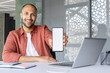 © Liubomir - Smiling man holding smartphone displaying screen at desk with laptop and notebook. Shows use of technology and business in modern office environment. Ideal for communication concept.