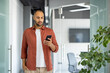© Liubomir - Young man in casual attire stands inside office using smartphone. Focused and connected, navigating workspace with phone in hand, exemplifying modern work style and communication.