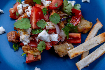 Salad with caramelized eggplant, cilantro, tomatoes and cream cheese on a blue plate with toasted bread on a gray background.