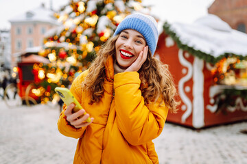 Young woman using a smartphone outside with the decorative christmas lights in the background. Сhristmas fair.