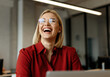 © Hanna - A woman in a red shirt laughs joyfully while sitting at her desk in a modern office during daytime
