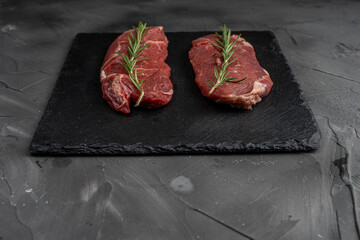 Two pieces of raw meat for steaks with a sprig of rosemary on a black mica board on a dark background.