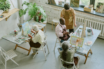 Wall Mural - High angle shot of senior students focused on painting while young adult teacher checking progress