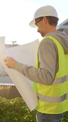 Male architect wearing safety hard hat and vest holding blueprint and looking to construction site at sunrise, back vertical view. Architecture and engineering