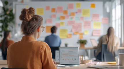 Creative brainstorming session in modern office with sticky notes on wall. woman in an orange sweater focuses on her laptop while colleagues collaborate in background