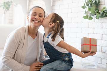 Wall Mural - Little small girl daughter giving present gift box making surprise to her mother on Birthday Christmas Mother`s day, hiding present behind , kissing her mom at home kitchen