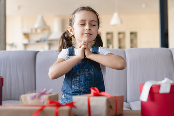 Wall Mural - Christmas wish to Santa Claus. Little girl daughter making a wish dreaming about new toys for birthday, sitting with eyes closed with present gift boxes at home