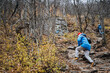 © Aleksey - A man, who is wearing a blue jacket, is skillfully climbing a challenging rocky path that winds its way through the dense woods, completely surrounded by the stunning beauty of nature all around him