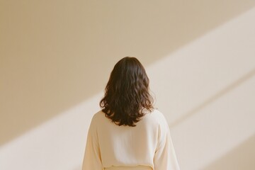 A woman in a stylish outfit stands gracefully against a pale taupe wall illuminated by soft natural light