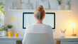 © Nano Photos - A person with tied-up hair works at a computer in a cozy home office, surrounded by plants, stationery, and warm lighting.