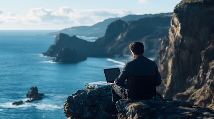 Wall Mural - A man with short dark hair working on his laptop from a caf perched on the edge of a cliff, with the ocean stretching out below him. The dramatic view and the fresh sea air add a sense of awe and