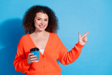 Poster - Photo of wavy haired model woman wearing orange cardigan directing finger empty space barista isolated on blue color background