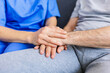 © Xavier Lorenzo - Close up view of mid adult female caregiver holding hands with senior patient sitting together on sofa at home. Elderly support and medical home assistance concept