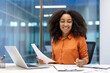 © Liubomir - Confident young businesswoman with documents smiling in modern office, engaged in work at her desk with laptop, pen, and paper, embodying professionalism and positivity