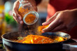 © Iryna - Hands adding vibrant orange spices into a hot pan while cooking in a kitchen