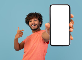 Cool handsome curly young indian guy in stylish pink t-shirt showing brand new cell phone with white empty screen and gesturing, sharing exciting online deal, blue background, mockup