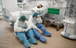 © My Ocean studio - Two exhausted healthcare workers in full protective gear sit on the floor beside a patient's hospital bed in an ICU, highlighting the emotional and physical toll of critical care in a medical setting.
