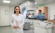 © My Ocean studio - Smiling female doctor stands with clipboard in front of linear accelerator in modern medical clinic, while patient receives radiotherapy treatment and another healthcare worker monitors procedure